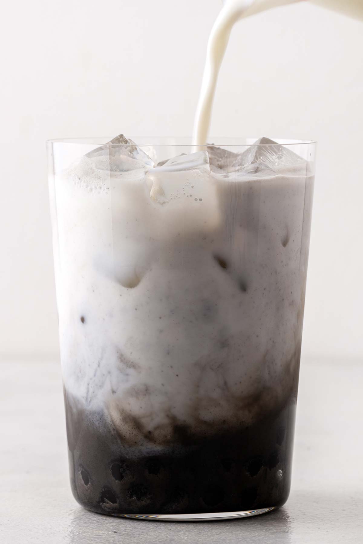 Milk being poured to make Black Sesame Bubble Tea (Black Sesame Boba Milk Tea) in a clear glass with wide straw.