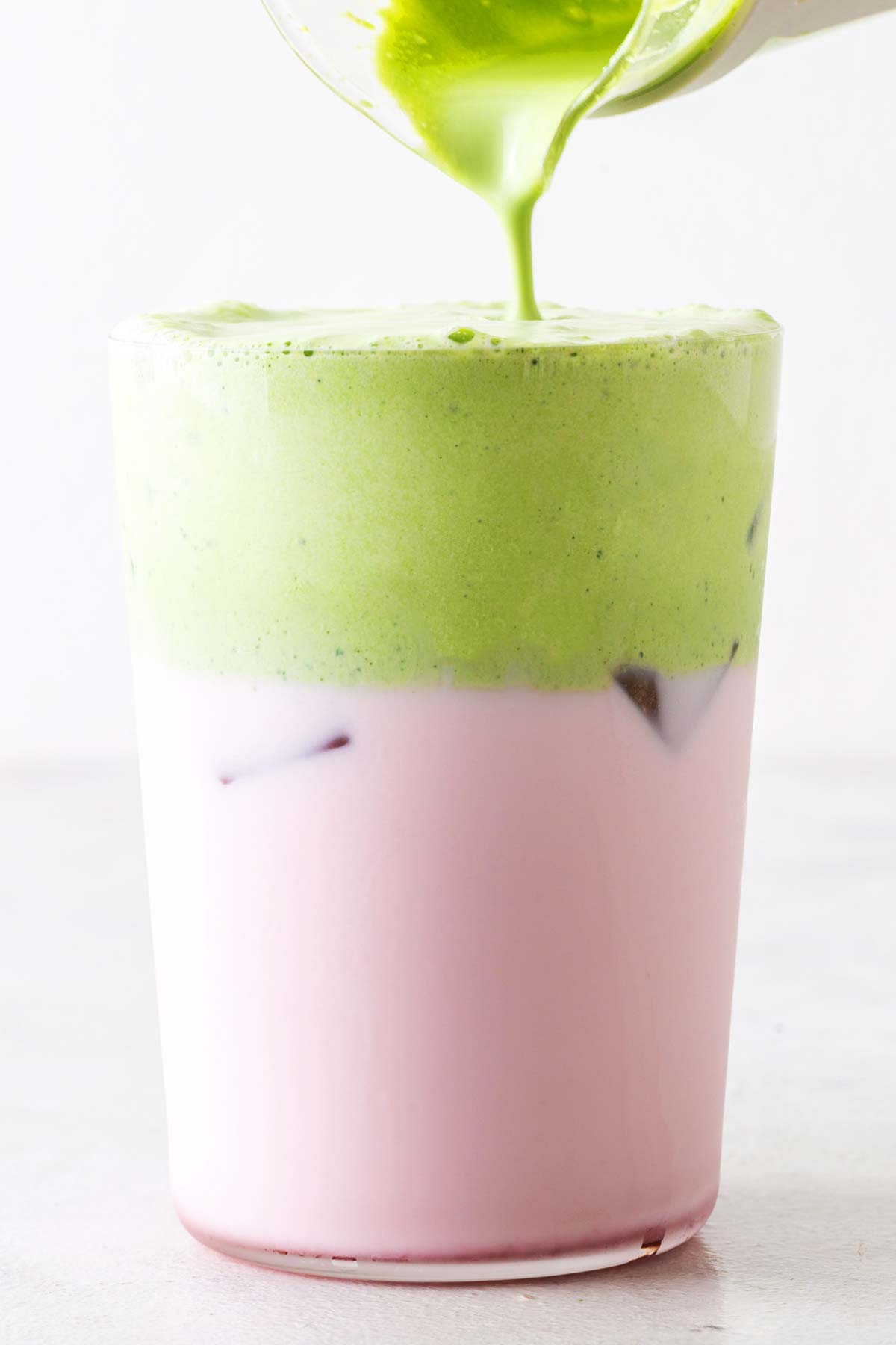 Matcha cold foam being poured over a pink iced drink in a clear glass.